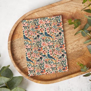 May include: A floral patterned notebook featuring a design of birds, pink flowers, and green leaves. The notebook is rectangular and rests on a wooden tray. The birds are blue and orange, set against a cream background.