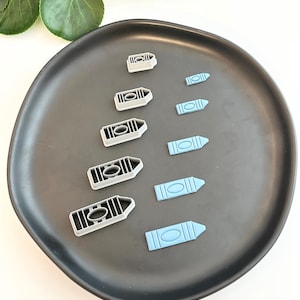 May include: Crayon-shaped cookie cutters in different sizes. The cutters are light blue and grey, arranged on a dark grey plate. The image shows a selection of baking tools.