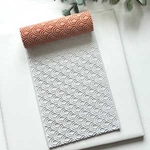 Könnte beinhalten: Eine weiße Teigrolle mit einem sich wiederholenden Muster aus überlappenden Halbkreisen. Die Teigrolle liegt auf einer weißen Oberfläche.