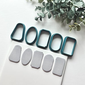 May include: A set of six teal plastic cookie cutters in various shapes, including a rectangle, oval, and a rounded rectangle. The cutters are on a white surface with six gray clay shapes that have been cut out using the cutters.