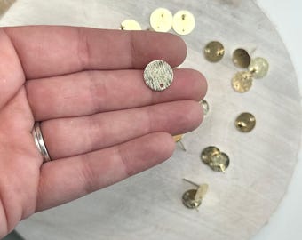 May include: A close-up of a gold-colored metal circle with a textured surface and a small hole in the center. The circle is being held by a hand, and there are several other similar circles scattered on a wooden surface.