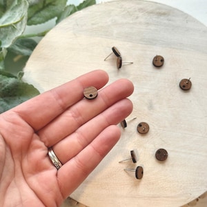 May include: A set of wooden circle earrings with a small hole in the center. The earrings are brown and have a natural wood grain. They are displayed on a wooden surface.