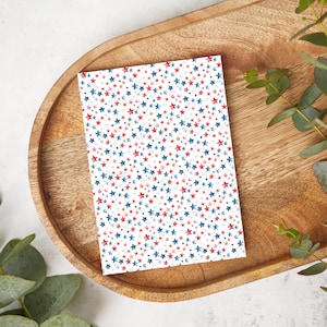 May include: A rectangular card with a white background covered in a pattern of red and blue stars. The card is placed on a wooden tray with a light brown color. Green leaves are in the background.