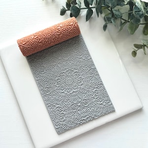May include: A brown patterned rolling pin rests on a gray, patterned sheet. The rolling pin and sheet are on a white surface, with green foliage in the background. The rolling pin and sheet have a floral design.