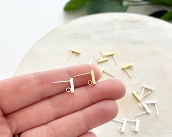 May include: A collection of earring posts in gold and silver tones. The posts have a rectangular bar design with a small loop at the bottom for attaching earring components. Some posts are held in a hand, while others are scattered on a light-colored surface.