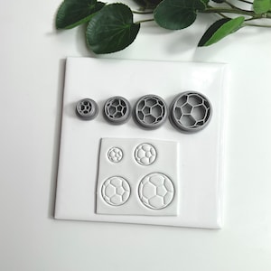 May include: A set of five gray plastic cookie cutters shaped like soccer balls. The cutters are arranged in order of size, with the largest cutter at the top and the smallest cutter at the bottom. The cutters are on a white surface. There is a white sheet of fondant with four soccer ball impressions on it.