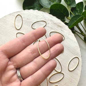 May include: A close-up of a hand holding a gold oval ring. The ring is made of metal and has a smooth finish. There are several other gold oval rings scattered on a white surface.