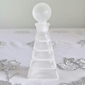 May include: A frosted glass bottle stopper with a spherical top and a tiered, conical body. The stopper is clear and sits on a white, patterned surface. The design is simple and elegant.