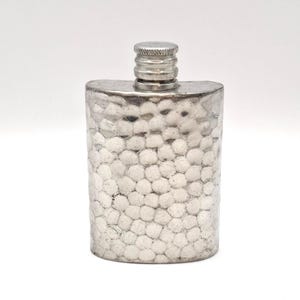 May include: A silver-coloured metal flask featuring a distinct hammered texture across its entire front surface. The container has a curved, rectangular body and a cylindrical, screw-top cap with horizontal ridges at the top centre of the vessel.