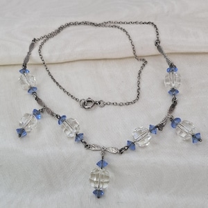 Op de afbeelding: Een delicate ketting met heldere, facetgeslepen kralen en kleine blauwe accenten. De ketting heeft een zilverkleurige ketting en een centrale hanger. Het ontwerp is symmetrisch, met de kralen gelijkmatig verdeeld.