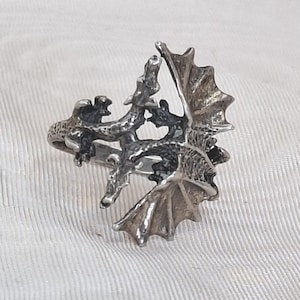May include: A silver-toned dragon ring. The ring features a detailed dragon design with outstretched wings and intricate scales. The dragon wraps around the finger, creating a unique and fantasy-inspired accessory.