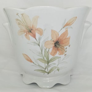 May include: White ceramic planter with a scalloped rim and two handles. The planter features a floral design with orange lilies and green leaves.