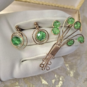 Peut inclure: Un ensemble de bijoux vintage composé d'une broche et d'une paire de boucles d'oreilles. La broche est en fil d'or et présente un groupe de pierres en verre vert. Les boucles d'oreilles sont également en fil d'or et ont chacune une seule pierre en verre vert.