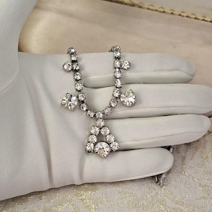 Op de afbeelding: Een zilveren ketting met een driehoekige hanger en meerdere kristallen accenten. De ketting ligt op een wit oppervlak.
