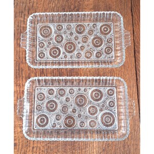 Puede incluir: Dos bandejas rectangulares de vidrio transparente con un patrón decorativo de círculos concéntricos. Cada bandeja tiene un borde festoneado y asas en los lados. Las bandejas se muestran sobre una superficie de madera.