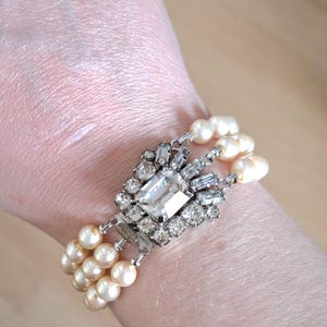 K&ouml;nnte beinhalten: Ein dreireihiges Perlenarmband mit einem gro&szlig;en, klaren, facettierten Strassstein als Mittelst&uuml;ck. Das Armband ist aus silberfarbenem Metall gefertigt und hat einen Verschluss.