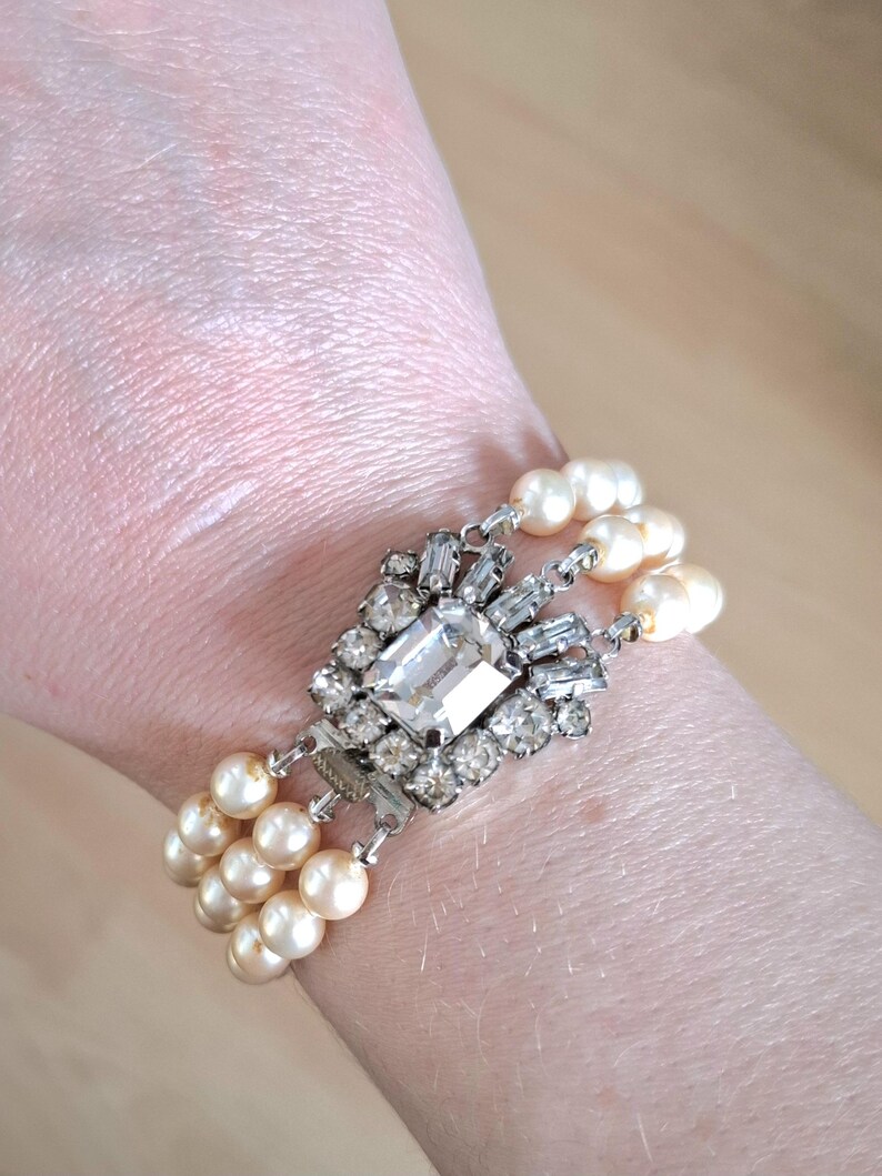 K&ouml;nnte beinhalten: Ein dreireihiges Perlenarmband mit einem gro&szlig;en, klaren, facettierten Strassstein als Mittelst&uuml;ck. Das Armband ist aus silberfarbenem Metall gefertigt und hat einen Verschluss.
