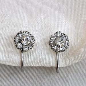 Op de afbeelding: Een paar zilverkleurige metalen clip-oorbellen, elk voorzien van een centrale heldere ronde strass-steen omringd door een cirkelvormige cluster van kleinere heldere strass-steentjes, wat een bloemachtig ontwerp met een gepolijste afwerking vormt.