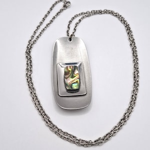 May include: A silver-toned metal pendant necklace featuring a rectangular dog tag style base with a brushed finish. Centered on the base is a smaller, raised square plate holding an iridescent, multi-colored abalone shell inlay piece.