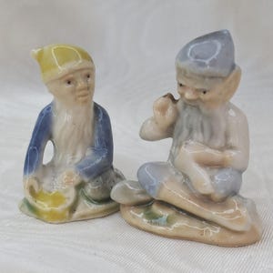May include: Two ceramic gnome figurines. One gnome wears a yellow hat and blue jacket, the other a blue hat and pants, smoking a pipe. Both have long white beards and are seated.