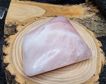 May include: A large, smooth, light pink rose quartz crystal with a white swirl pattern. The crystal is shaped like a triangle and is sitting on a wooden surface.
