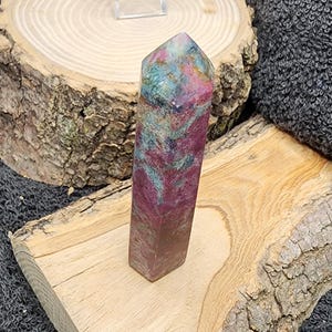 May include: A tall, rectangular crystal with a pointed top. The crystal features a blend of colors, including red, green, and purple. It rests on a piece of wood, with a wooden slice and dark gray fabric in the background.