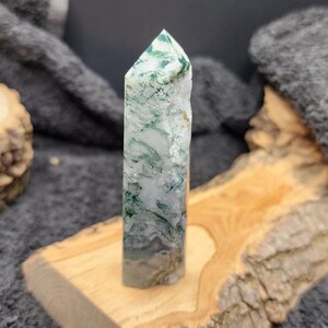May include: A green and white agate crystal point, also known as a wand, is displayed on a wooden stand. The crystal has a unique swirling pattern of green and white.