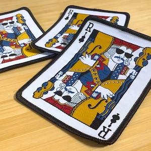Op de afbeelding: Drie speelkaartenpatches met een cartoonkoning met een zonnebril en een muziekinstrument. De kaarten hebben een witte achtergrond, een zwarte rand en de letter "R" in de hoeken. De kleding van de koning omvat blauw, geel en rood.