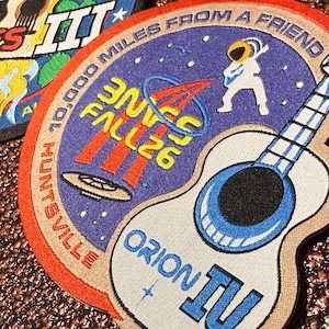 May include: A circular embroidered patch featuring a purple space scene with a white astronaut, a flying saucer, and a blue acoustic guitar. The text reads 10,000 MILES FROM A FRIEND, HUNTSVILLE, UNITE FALL 26, and ORION IV in yellow, red, and blue.