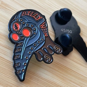 Könnte beinhalten: Eine schwarze und braune Metallnadel mit einem detaillierten Design eines Vogelkopfes, der eine Gasmaske trägt. Die Nadel trägt die Worte "Billy Ween" und die Nummer "45/50" auf der Rückseite.