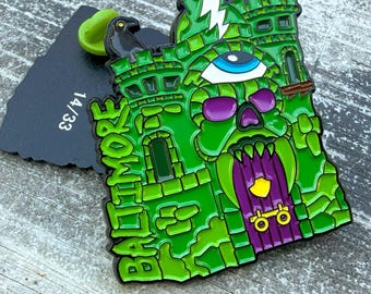 May include: A green enamel pin shaped like a castle with a skull, a single eye, and a black raven on top. The text BALTIMORE is written vertically on the left side. The back of the pin is black with the text 14/33 printed in white.