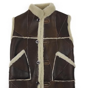 Pode incluir: Um colete de couro castanho com um design de patchwork. O colete apresenta forro e acabamento em pele de carneiro creme ao redor da gola, frente e bolsos. Possui fecho de botão e dois bolsos frontais.
