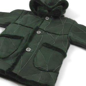 Könnte beinhalten: Eine dunkelgrüne Kapuzenjacke aus Schaffell mit Patchwork-Muster und Knopfleiste. Die Jacke hat einen Pelzbesatz an der Kapuze und den Taschen.