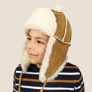 May include: A brown faux fur aviator hat with a white faux fur lining and ear flaps. The hat has a brown leather strap with a pom pom on top.