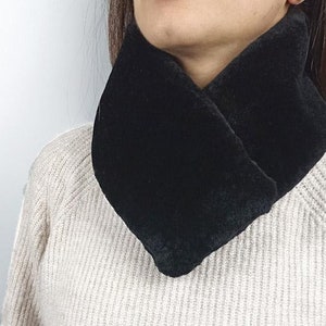 Puede incluir: Un calentador de cuello de piel sintética negra. El calentador de cuello está doblado por la mitad y se lleva alrededor del cuello. El calentador de cuello está hecho de piel sintética suave y mullida.