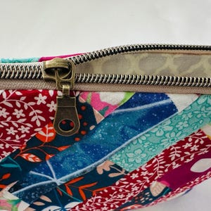 K&ouml;nnte beinhalten: Nahaufnahme eines farbenfrohen Patchwork-Stoffbeutels mit einem bronzefarbenen Rei&szlig;verschluss. Der Stoff weist eine Vielzahl von Mustern und Farben auf, darunter Rot, Blau und Rosa. Der Rei&szlig;verschluss ist teilweise ge&ouml;ffnet.