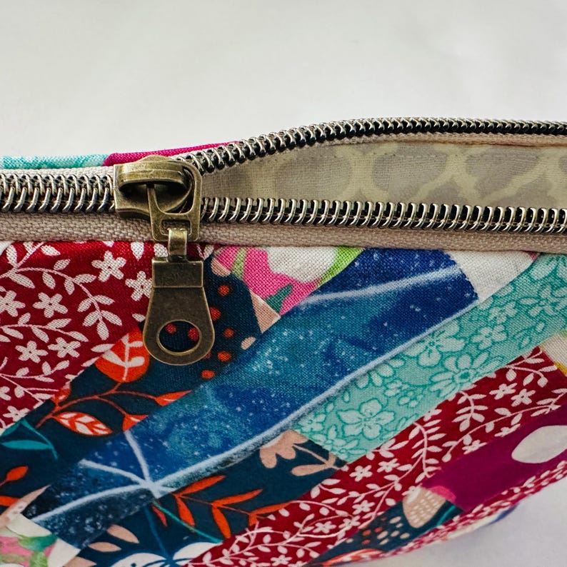 K&ouml;nnte beinhalten: Nahaufnahme eines farbenfrohen Patchwork-Stoffbeutels mit einem bronzefarbenen Rei&szlig;verschluss. Der Stoff weist eine Vielzahl von Mustern und Farben auf, darunter Rot, Blau und Rosa. Der Rei&szlig;verschluss ist teilweise ge&ouml;ffnet.
