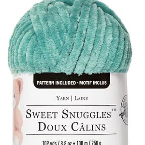 Puede incluir: Una madeja de hilo Sweet Snuggles turquesa. La etiqueta dice "Patrón incluido" y "Sweet Snuggles Doux Câlins". El hilo es de 100 m / 250 g. El hilo es un material suave y afelpado, perfecto para proyectos de punto o ganchillo.