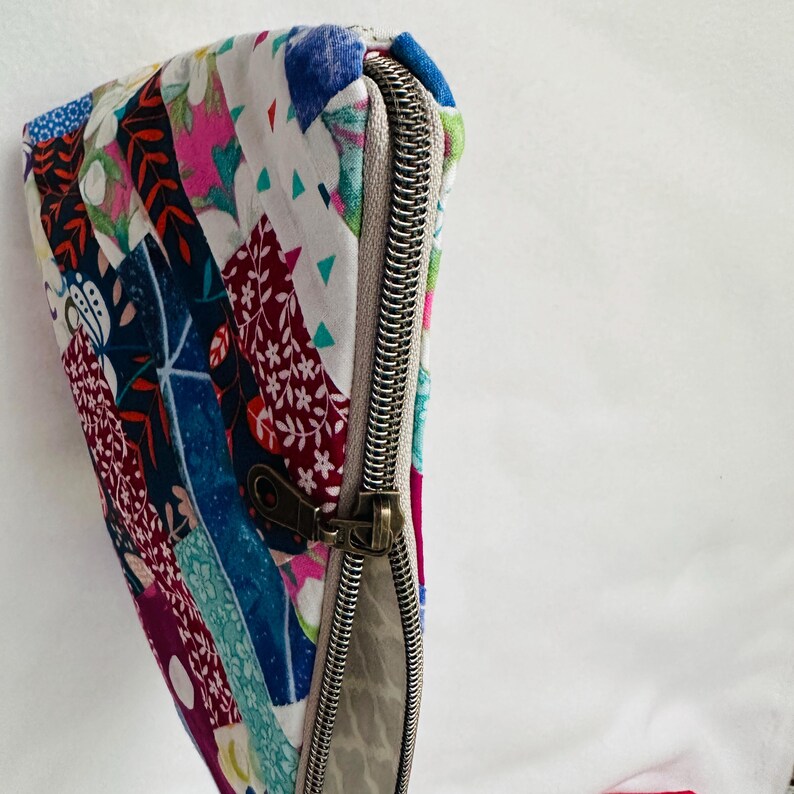 K&ouml;nnte beinhalten: Eine bunte Patchwork-Rei&szlig;verschlusstasche. Sie zeigt verschiedene Stoffmuster, darunter florale, geometrische und abstrakte Designs in Blau-, Rot-, Rosa- und Wei&szlig;t&ouml;nen. Der Rei&szlig;verschluss ist silberfarben, der Zipper bronzefarben.