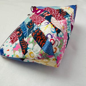 K&ouml;nnte beinhalten: Ein farbenfrohes, gestepptes T&auml;schchen mit Rei&szlig;verschluss. Das Patchwork-Design zeigt verschiedene Blumen- und geometrische Muster in Blau-, Rot-, Rosa- und Gelbt&ouml;nen. Das T&auml;schchen hat eine dreieckige Form und scheint handgefertigt zu sein.
