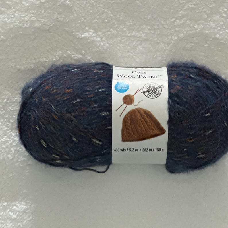 May include: A skein of blue yarn with flecks of white and brown. The yarn is labeled "Cozy Wool Tweed" and is 418 yards / 5.2 ounces / 382 meters / 150 grams.