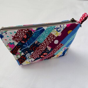 K&ouml;nnte beinhalten: Handgefertigte Rei&szlig;verschlusstasche mit Patchwork-Design. Die Tasche besteht aus Stoffstreifen in verschiedenen Farben, darunter Blau, Rot und Pink. Eine kleine Schlaufe aus rot-wei&szlig; gemustertem Stoff ist am Rei&szlig;verschluss befestigt.