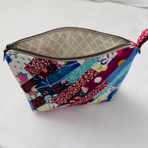 K&ouml;nnte beinhalten: Ein farbenfrohes, offenes T&auml;schchen mit Rei&szlig;verschluss und Patchwork-Design. Die Au&szlig;enseite zeigt Stoffstreifen in verschiedenen Farben und Mustern, darunter Blumen- und geometrische Drucke. Das Innenfutter ist hellbeige mit einem sich wiederholenden Muster. Eine kleine rote Schlaufe ist am Rei&szlig;verschluss befestigt.