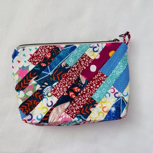 K&ouml;nnte beinhalten: Ein farbenfrohes, gestepptes T&auml;schchen mit Rei&szlig;verschluss. Es besteht aus Stoffstreifen in verschiedenen Mustern und Farben, darunter Blumen- und geometrische Designs. Der Rei&szlig;verschluss hat eine kleine, geknotete Kordel.