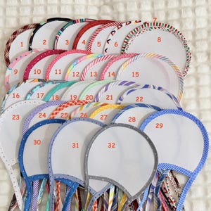 May include: A collection of thirty-two circular fabric headbands with white centers and various patterned borders, each labeled with a red number from one to thirty-two. The headbands feature diverse colors and prints, including stripes, dots, and plaids.