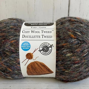 Puede incluir: Un ovillo de lana Loops & Threads Cozy Wool Tweed en el color Douillette Tweed. La lana es una mezcla de lana y acrílico y tiene una textura de tweed. El ovillo está etiquetado con el nombre de la lana y el color, así como el nombre de la marca. La etiqueta también incluye un patrón para un gorro.