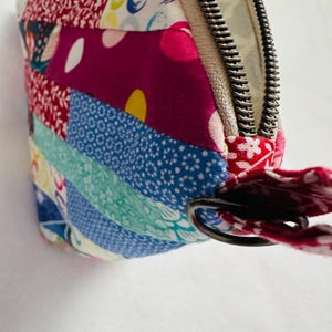 K&ouml;nnte beinhalten: Nahaufnahme eines farbenfrohen Patchwork-Rei&szlig;verschlussetuis. Das Etui weist eine Vielzahl von gemusterten Stoffen in Rot-, Blau-, Rosa- und Wei&szlig;t&ouml;nen auf. Ein silberner Rei&szlig;verschluss und eine rote Stoffschlaufe sind sichtbar.
