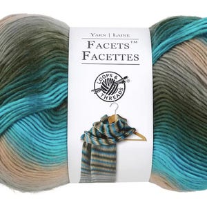 Puede incluir: Una madeja de hilo Facets de Loops & Threads en un degradado de color turquesa, verde y beige. El hilo está envuelto alrededor de una etiqueta blanca con el texto "Yarn | Laine Facets™ Facettes" y un logotipo de Loops & Threads.