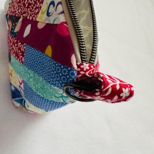 K&ouml;nnte beinhalten: Ein Patchwork-Etui mit Rei&szlig;verschluss und einer Vielzahl farbenfroher Stoffmuster. Das Etui hat einen silbernen Rei&szlig;verschluss und eine rote Stoffschlaufe mit wei&szlig;em Blumendesign. Das Etui besteht aus verschiedenen Stoffen in Rot-, Blau- und Lilat&ouml;nen.