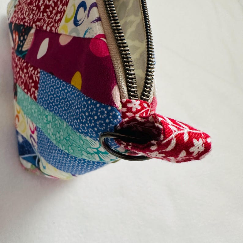 K&ouml;nnte beinhalten: Ein Patchwork-Etui mit Rei&szlig;verschluss und einer Vielzahl farbenfroher Stoffmuster. Das Etui hat einen silbernen Rei&szlig;verschluss und eine rote Stoffschlaufe mit wei&szlig;em Blumendesign. Das Etui besteht aus verschiedenen Stoffen in Rot-, Blau- und Lilat&ouml;nen.